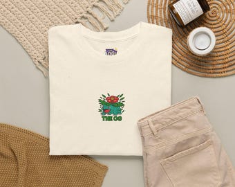 Puede incluir: Una camiseta de color crema con un pequeño bordado que representa una criatura verde con una flor roja en la espalda, acompañada del texto verde "THE OG" debajo. La camiseta tiene un cuello redondo y una pequeña etiqueta de marca interior.