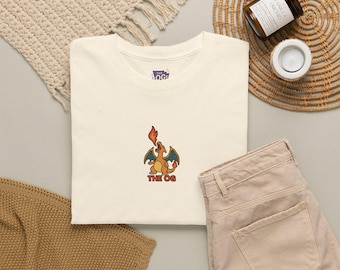 May include: An off-white crew neck t-shirt featuring an embroidered graphic of a small orange dragon breathing fire with the text THE OG underneath. The shirt has a small interior label with the text THE OGs printed in purple and yellow.