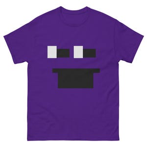 Puede incluir: Una camiseta morada con un diseño de cara pixelada. El diseño incluye dos cuadrados blancos para los ojos y un rectángulo negro para la boca. La camiseta está hecha de un material suave y tiene mangas cortas.