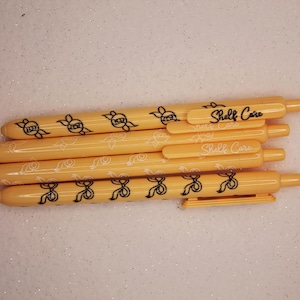 Puede incluir: Una colección de bolígrafos amarillos con diseños de lazos y cintas negras. Los bolígrafos tienen el texto "Shelf Care" impreso. Los bolígrafos están dispuestos en una pila diagonal sobre una superficie blanca y brillante.