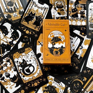 Puede incluir: Un juego de cartas de tarot con una caja naranja central. La caja dice "Adorable Cat Tarot 2024". Las cartas son negras y doradas, con ilustraciones de gatos, lunas y estrellas. Las cartas están esparcidas alrededor de la caja.