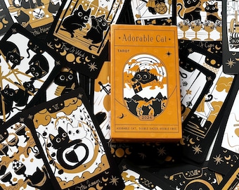 Könnte beinhalten: Ein Tarotkartenspiel mit einer orangefarbenen Box in der Mitte. Auf der Box steht "Adorable Cat Tarot 2024". Die Karten sind schwarz und goldfarben und zeigen Katzenillustrationen, Monde und Sterne. Die Karten sind um die Box herum verstreut.