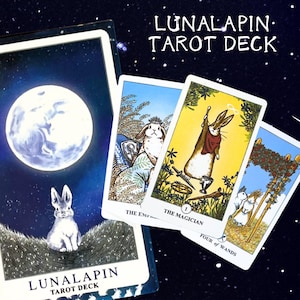 Könnte beinhalten: Ein Tarotdeck mit dem Titel "LUNALAPIN TAROT DECK" wird mit mehreren Karten gezeigt. Die Karten zeigen Illustrationen von Kaninchen in verschiedenen Szenen, darunter eine mit Vollmond und einem Kaninchen auf einem Feld. Die Karten sind vor einem Sternenhintergrund angeordnet.