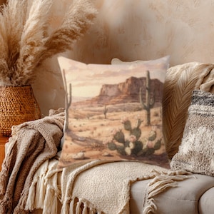 May include: Square decorative pillow featuring a desert landscape with cacti, mountains, and a warm color palette. The pillow is placed on a beige sofa with a fringed throw blanket, creating a cozy, Southwestern-inspired aesthetic.