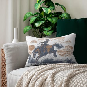 Pode incluir: Almofada retangular com um cowboy montando um cavalo selvagem. O design apresenta uma ilustração marrom e azul de um cowboy laçando um cavalo, em um cenário de paisagem desértica. A almofada está em um sofá cinza com um cobertor creme.