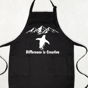 May include: Black apron with two front pockets and long ties. The apron features a white graphic of a penguin in front of a mountain range, with the text "Difference is Creative" below the penguin.