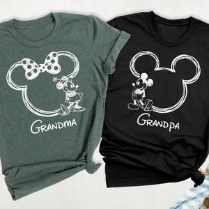May include: Two t-shirts, one dark green and one black, each featuring a white outline of Mickey Mouse's head. The dark green shirt has a bow and the word "Grandma" below. The black shirt has the word "Grandpa".