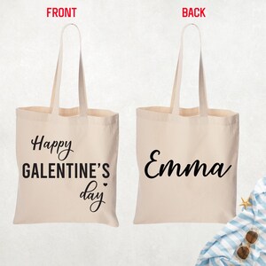 May include: A beige canvas tote bag with long handles. The front of the bag says "Happy Galentine's Day" in black script. The back of the bag has the name "Emma" in black script.