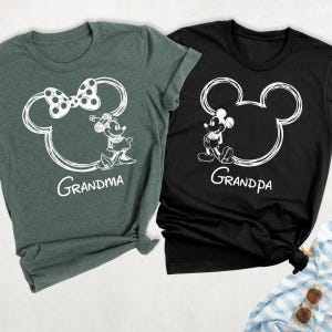 May include: Two t-shirts, one dark green and one black, each featuring a white outline of Mickey Mouse's head. The dark green shirt has a bow and the word "Grandma", while the black shirt says "Grandpa".