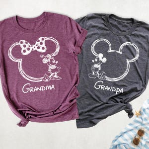 May include: Two t-shirts, one burgundy and one charcoal gray, featuring white cartoon character outlines. The burgundy shirt has a Minnie Mouse design with the word "Grandma." The gray shirt has a Mickey Mouse design with the word "Grandpa."