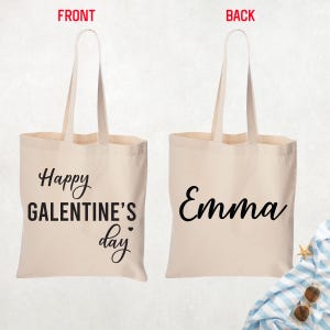 May include: A beige canvas tote bag with long handles. The front of the bag says "Happy Galentine's Day" in black script. The back of the bag has the name "Emma" in black script.