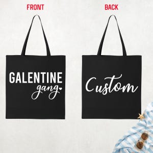 May include: Black tote bag with white text. The front of the bag says "GALENTINE gang" with a heart. The back of the bag says "Custom" in a cursive font. The bag has two black handles.