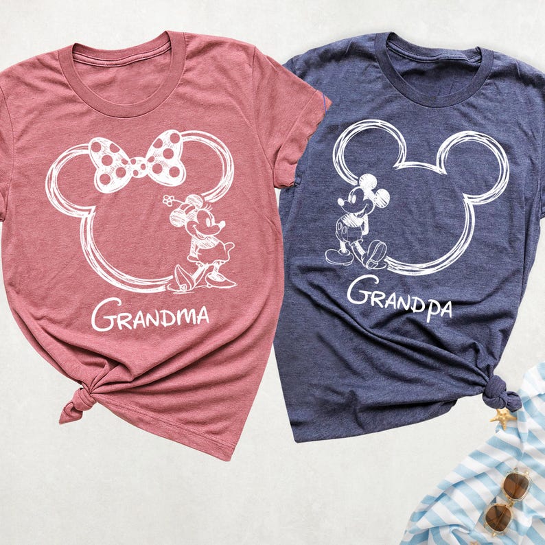 May include: Two t-shirts, one mauve and one navy blue, featuring white Mickey and Minnie Mouse designs. The mauve shirt says "Grandma" and the navy shirt says "Grandpa". Both shirts are knotted at the bottom.