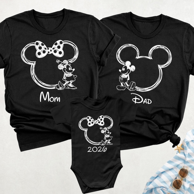 May include: Black family matching t-shirts and a onesie featuring a white Mickey Mouse outline with Minnie Mouse and Mickey Mouse characters. The shirts have the words "Mom" and "Dad", and the onesie has the number "2026".