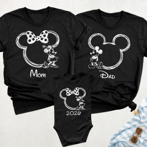 May include: Black family matching t-shirts and a onesie featuring a white Mickey Mouse outline with Minnie Mouse and Mickey Mouse characters. The shirts have the words "Mom" and "Dad", and the onesie has the number "2026".