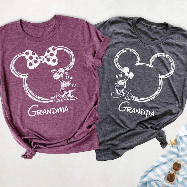 May include: Two t-shirts, one burgundy and one charcoal gray, featuring white cartoon illustrations. The burgundy shirt has a Minnie Mouse design with the word "Grandma" below. The gray shirt has a Mickey Mouse design with the word "Grandpa" below. Both shirts are short-sleeved.