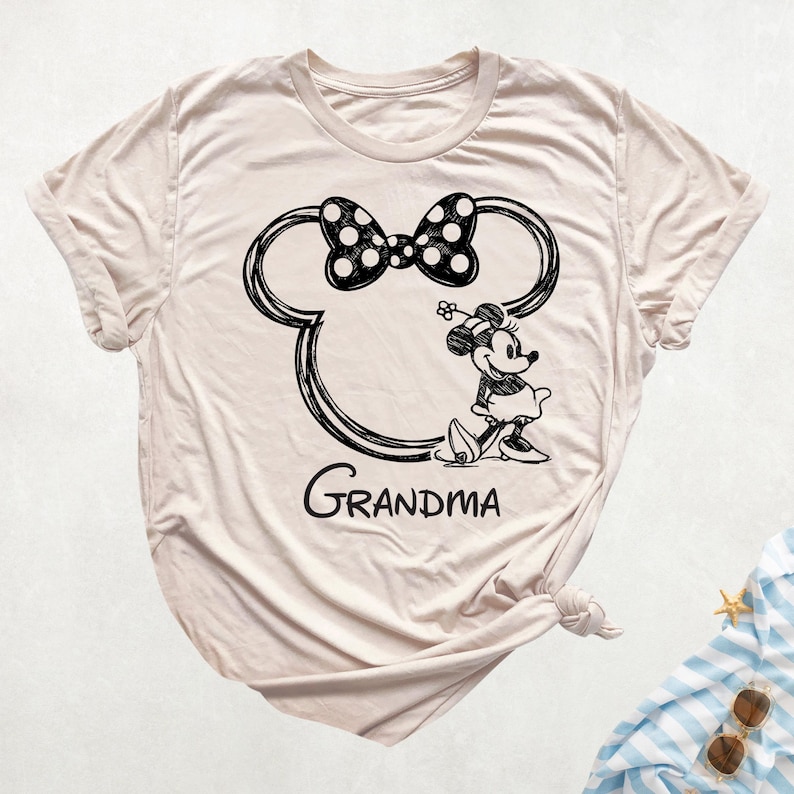 May include: A light beige t-shirt featuring a black graphic of Minnie Mouse's head with a polka dot bow and the word "Grandma" below. Minnie Mouse is depicted in a walking pose, wearing a dress and hat. The shirt is knotted at the bottom.