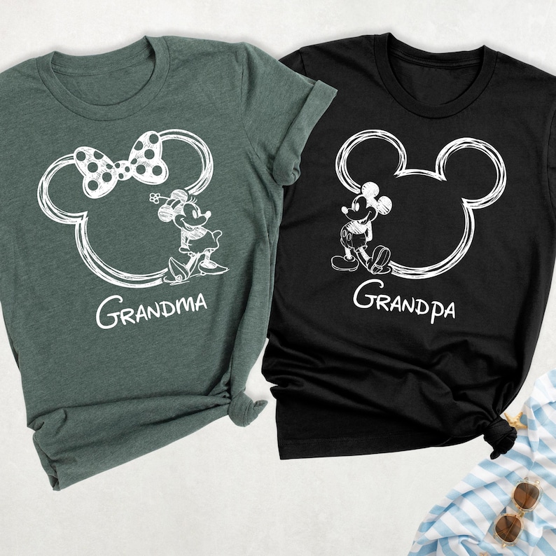 May include: Two t-shirts, one dark green and one black, each featuring a white outline of Mickey Mouse's head. The dark green shirt has a bow and the word "Grandma" below. The black shirt has the word "Grandpa".