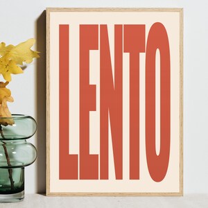Może przedstawiać: Oprawiony wydruk artystyczny ze słowem "LENTO" w dużych, pogrubionych, rdzawych literach na kremowym tle. Grafika jest w drewnianej ramie i jest eksponowana w pobliżu szklanego wazonu z kilkoma żółtymi liśćmi.