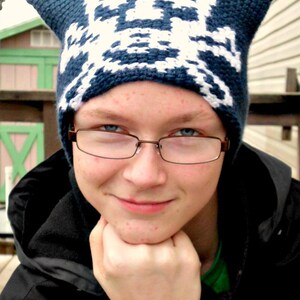 May include: A person wearing a blue and white knit hat with cat ears and brown framed glasses. They are wearing a black jacket and have a serious expression.
