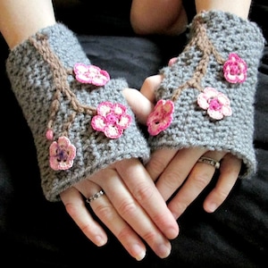 May include: A pair of gray crocheted fingerless gloves with pink and white crocheted flowers. The flowers are arranged in a pattern that resembles a branch with blossoms.