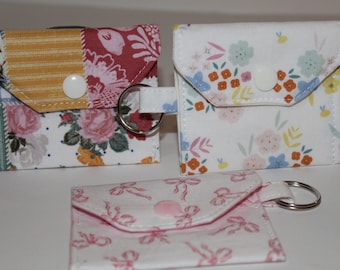 May include: Three small, square fabric pouches with key rings. Two pouches have floral patterns, one with pink roses and the other with colorful flowers. The third pouch has a white background with pink bows.