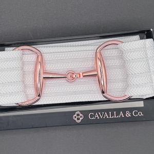 May include: A light gray horse bit with rose gold accents, presented in a black box. The bit is part of a wider, elasticated strap. The box features the text "CAVALLA & Co." and a logo.