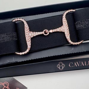 May include: Black elastic belt with rose gold-colored hardware, including a buckle and a D-ring. The buckle is embellished with small, sparkling crystals. The belt is presented in a black box with the brand name "Cavalla & Co."