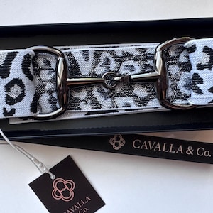 May include: A black and white leopard print belt with silver-tone hardware, presented in a black box. The belt features a classic equestrian-style buckle. A tag with the brand name "Cavalla & Co." is attached.