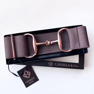 May include: A wide, dark brown elastic belt featuring a rose gold-toned metal bit-style buckle. The belt is presented in a black rectangular box labeled CAVALLA & Co. and includes a matching black hang tag with the brand logo and name printed in rose gold.
