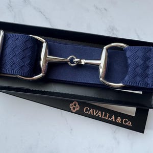 May include: A navy blue elastic belt featuring a silver-toned metal horse bit buckle with a textured woven pattern on the ends. The belt is presented in a black rectangular box with the text CAVALLA & Co. printed in a metallic copper color.
