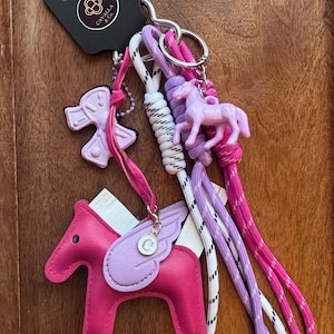 May include: A collection of keychains featuring horse and equestrian-themed charms. The keychains include a pink leather horse with wings, a light pink plastic horse, and a light pink bow-shaped charm. The keychains are attached to a metal ring with pink, white, and purple cords.