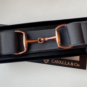 May include: A dark gray elastic belt featuring a rose gold-toned metal horse bit buckle. The belt is presented in a black rectangular box with the text CAVALLA & Co. printed in gold on the front of the packaging, showcasing a sleek design.