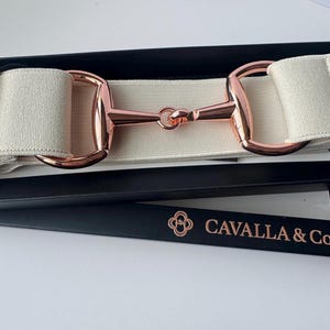 May include: An elastic belt in a cream color featuring a rose gold horse bit buckle. The belt is presented in a black rectangular box with the text CAVALLA & Co. printed in rose gold on the lid. The design includes a metal loop on one side.