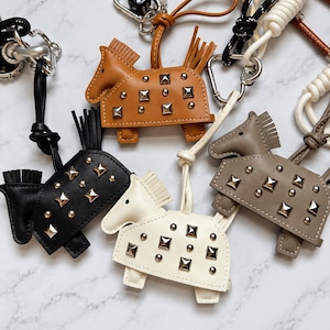 May include: A collection of horse-shaped bag charms crafted from leather in black, tan, white, and taupe. Each charm features metallic pyramid studs, a fringed mane and tail, and is attached to a knotted cord with a silver-toned metal clasp.