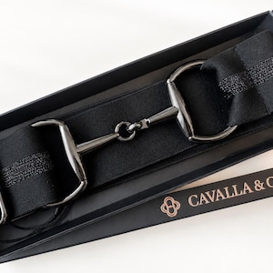 May include: A black elastic belt featuring a gunmetal-toned horse bit buckle with a central hinge. The belt strap has a subtle shimmering horizontal stripe. It is presented in a black rectangular box labeled with the text CAVALLA & Co.