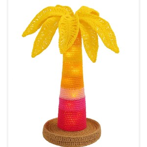 Pode incluir: Uma escultura de palmeira de crochê feita à mão. A árvore apresenta uma copa amarela, um tronco multicolorido em tons de amarelo, laranja, rosa e vermelho, e assenta sobre uma base circular castanha. É um objeto decorativo.