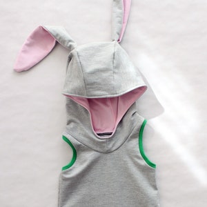 Puede incluir: Un vestido gris sin mangas con capucha, orejas de conejo rosas, ribetes verdes y detalles a rayas blancas y negras.