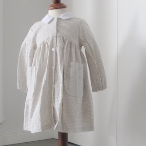 Könnte beinhalten: Ein helles, langärmliges Kleid mit weißem Kragen und zwei Vordertaschen wird auf einer Holzpuppe präsentiert. Das Kleid hat ein dezentes Karomuster und eine Knopfleiste vorne. Die Puppe steht auf einem runden Holzsockel.