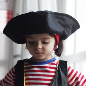 May include: A child dressed in a pirate costume. The outfit includes a black tricorn hat, a red and white striped long-sleeved shirt, a black vest with gold trim, and a red bandana. The vest has a decorative white bow and a blue and gold accent.