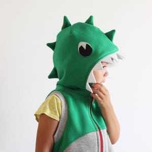 Puede incluir: Un ni&ntilde;o lleva un chaleco con capucha de dinosaurio verde y gris con una cremallera blanca. La capucha tiene una boca blanca con dientes y un ojo negro.