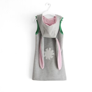 Puede incluir: Un vestido gris con capucha, orejas de conejo rosas y una flor gris en la espalda. El vestido tiene un ribete verde alrededor del cuello y las sisas.