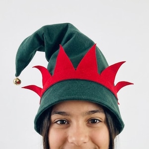 Peut inclure: Un chapeau d'elfe vert avec une bordure rouge et une petite cloche dorée. Le chapeau est porté par une personne portant un pull en tricot torsadé crème. Le fond est blanc uni.