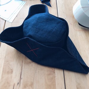 Pode incluir: Um chap&eacute;u tricornio de ganga azul escuro com um detalhe de ponto de cruz vermelho. O chap&eacute;u est&aacute; numa superf&iacute;cie de madeira, com retalhos de tecido e uma m&aacute;quina de costura vis&iacute;veis no fundo. Acess&oacute;rio de figurino hist&oacute;rico.