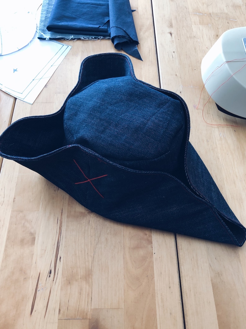 Pode incluir: Um chap&eacute;u tricornio de ganga azul escuro com um detalhe de ponto de cruz vermelho. O chap&eacute;u est&aacute; numa superf&iacute;cie de madeira, com retalhos de tecido e uma m&aacute;quina de costura vis&iacute;veis no fundo. Acess&oacute;rio de figurino hist&oacute;rico.