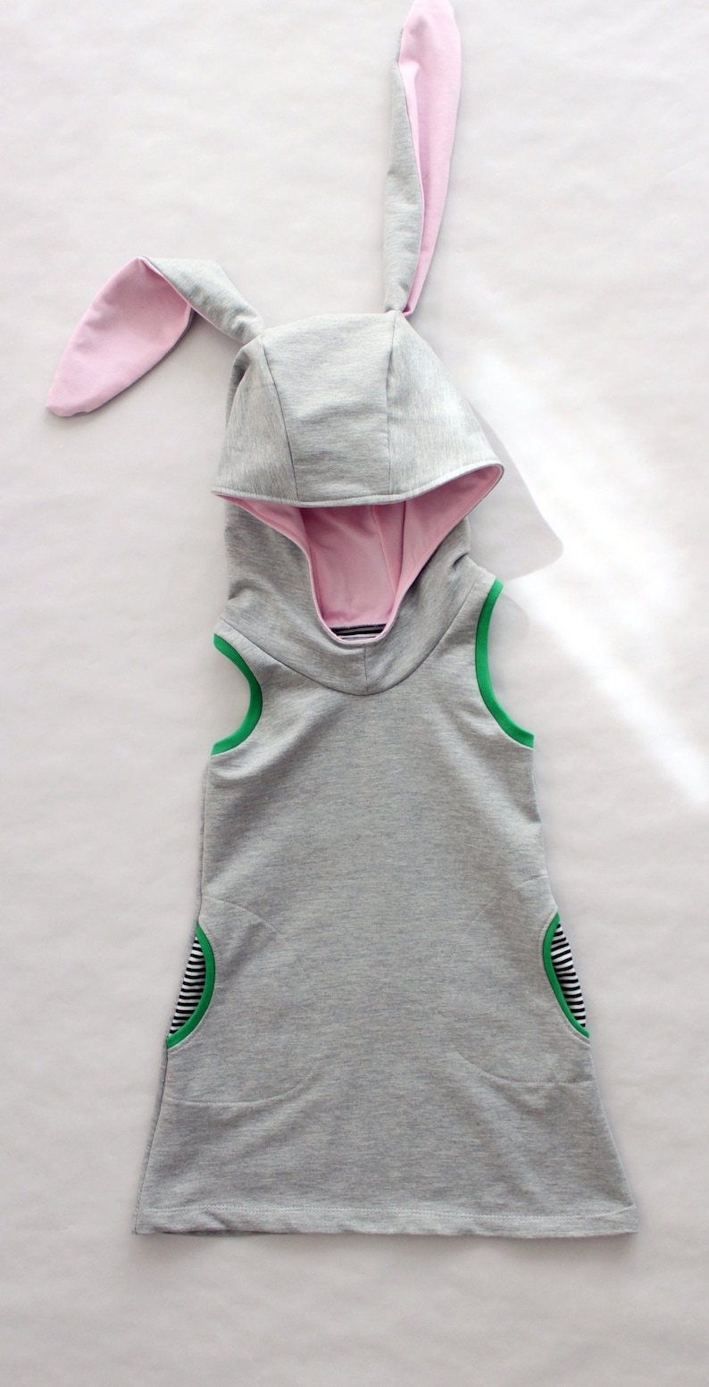 Puede incluir: Un vestido gris sin mangas con capucha, orejas de conejo rosas, ribetes verdes y detalles a rayas blancas y negras.