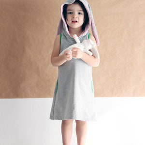 Puede incluir: Una ni&ntilde;a peque&ntilde;a con un vestido gris con capucha y orejas de conejo rosas. El vestido tiene ribetes verdes y la ni&ntilde;a sostiene un conejito de peluche blanco.