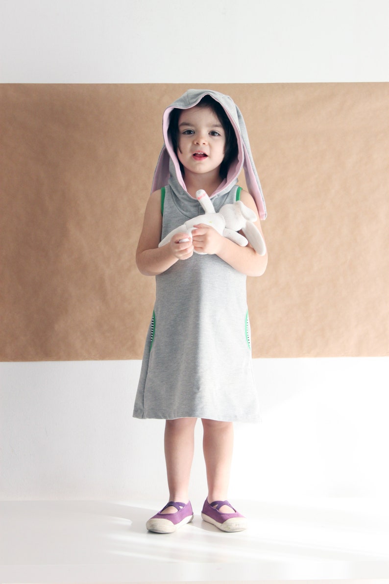 Puede incluir: Una ni&ntilde;a peque&ntilde;a con un vestido gris con capucha y orejas de conejo rosas. El vestido tiene ribetes verdes y la ni&ntilde;a sostiene un conejito de peluche blanco.