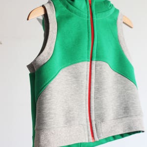 Puede incluir: Un chaleco sin mangas con capucha y una cremallera roja. El chaleco est&aacute; hecho de dos telas diferentes: una tela verde en la parte superior y una tela gris en la parte inferior.