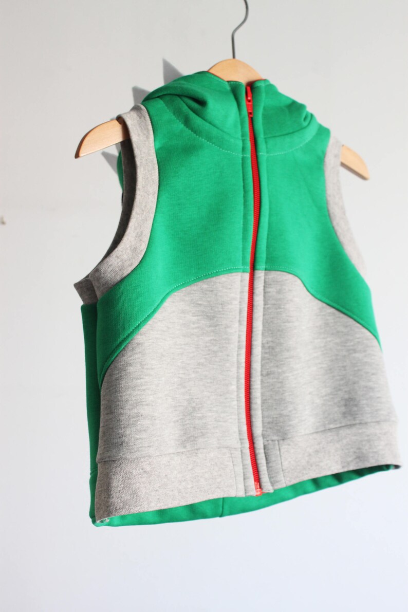 Puede incluir: Un chaleco sin mangas con capucha y una cremallera roja. El chaleco est&aacute; hecho de dos telas diferentes: una tela verde en la parte superior y una tela gris en la parte inferior.
